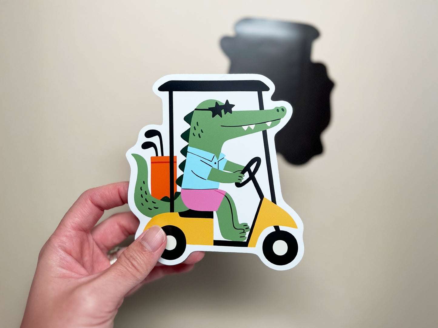 Hand holding funny die-cut magnet of cartoon alligator in golf cart with clubs, quirky novelty fridge magnet gift for golfers and golf lovers."