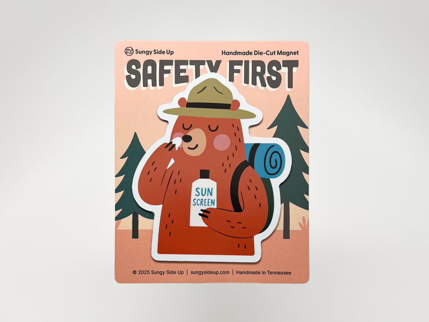 Handmade die-cut fridge magnet of a cartoon bear camper packaged on a peach “SAFETY FIRST” backing card. The brown bear wears a ranger hat, carries a blue backpack, and applies sunscreen from a bottle. Quirky camping magnet gift for hikers, national park fans, and outdoor adventurers.