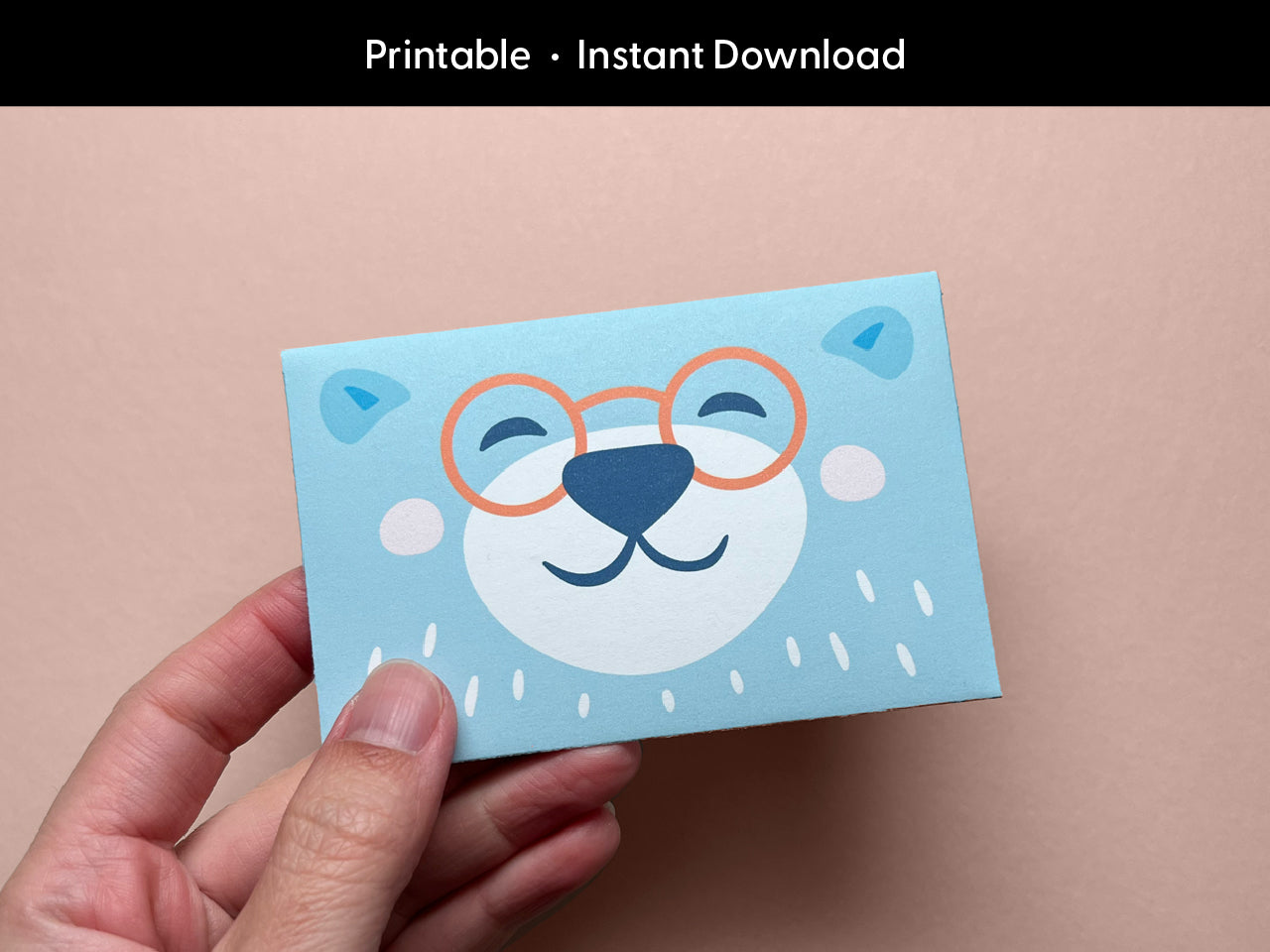 A hand holding a cute printable bear gift card holder featuring a smiling bear face with orange glasses on a light blue background. The bear illustration is cheerful and whimsical, perfect for kids' birthdays, baby showers, or teacher appreciation gifts. This digital download is easy to print and assemble at home, making your gift card presentation extra special and fun for any celebration or occasion.