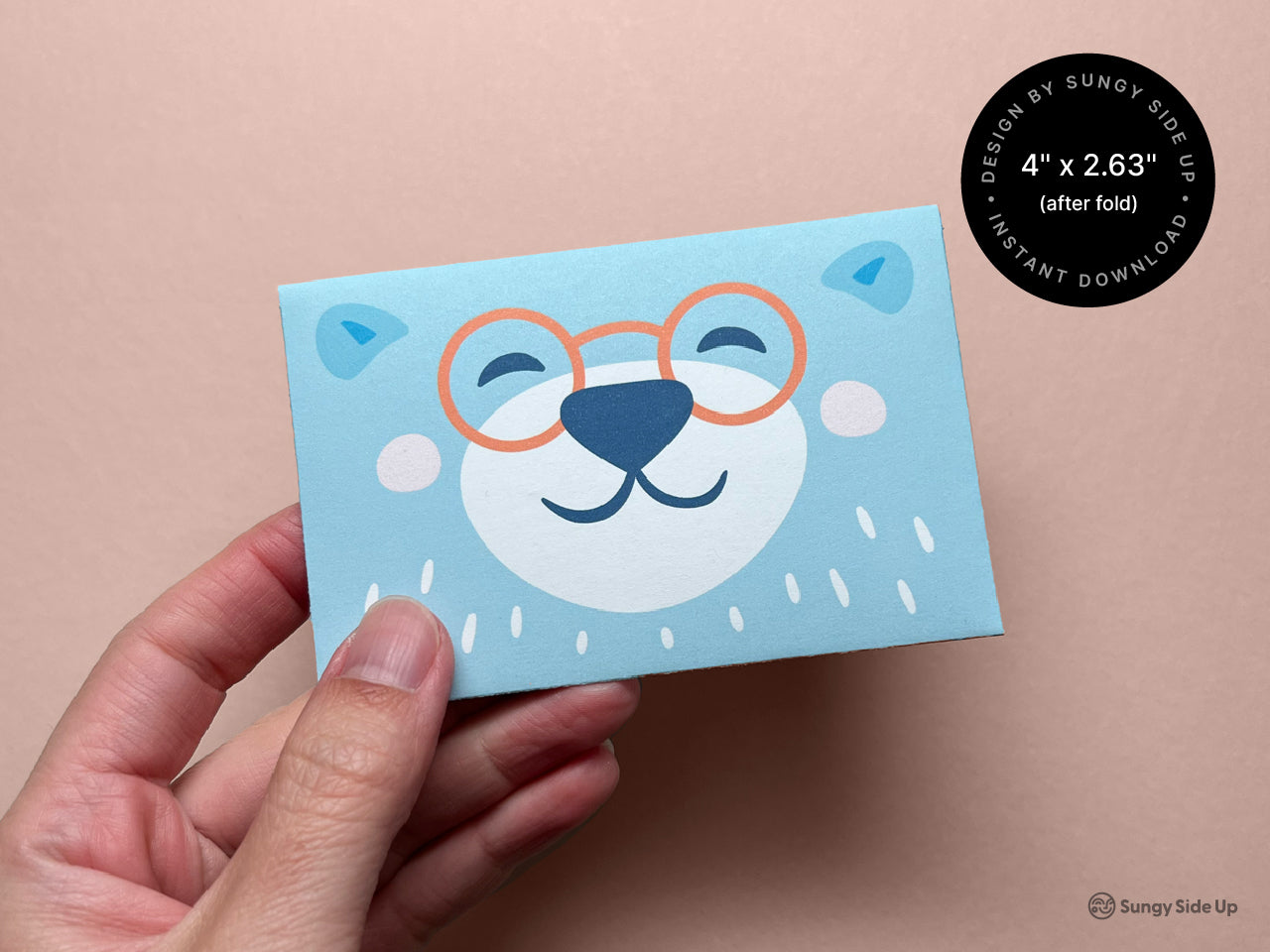 Front view of the finished printable bear gift card holder held in hand, showing a cheerful bear face with orange glasses on a blue background. This compact 4 x 2.63 inch design is perfect for dressing up a gift card with personality. Ideal for kids, teachers, or anyone who loves cute animals. A fun, easy DIY project that you can print and assemble at home for thoughtful gifting on any occasion.