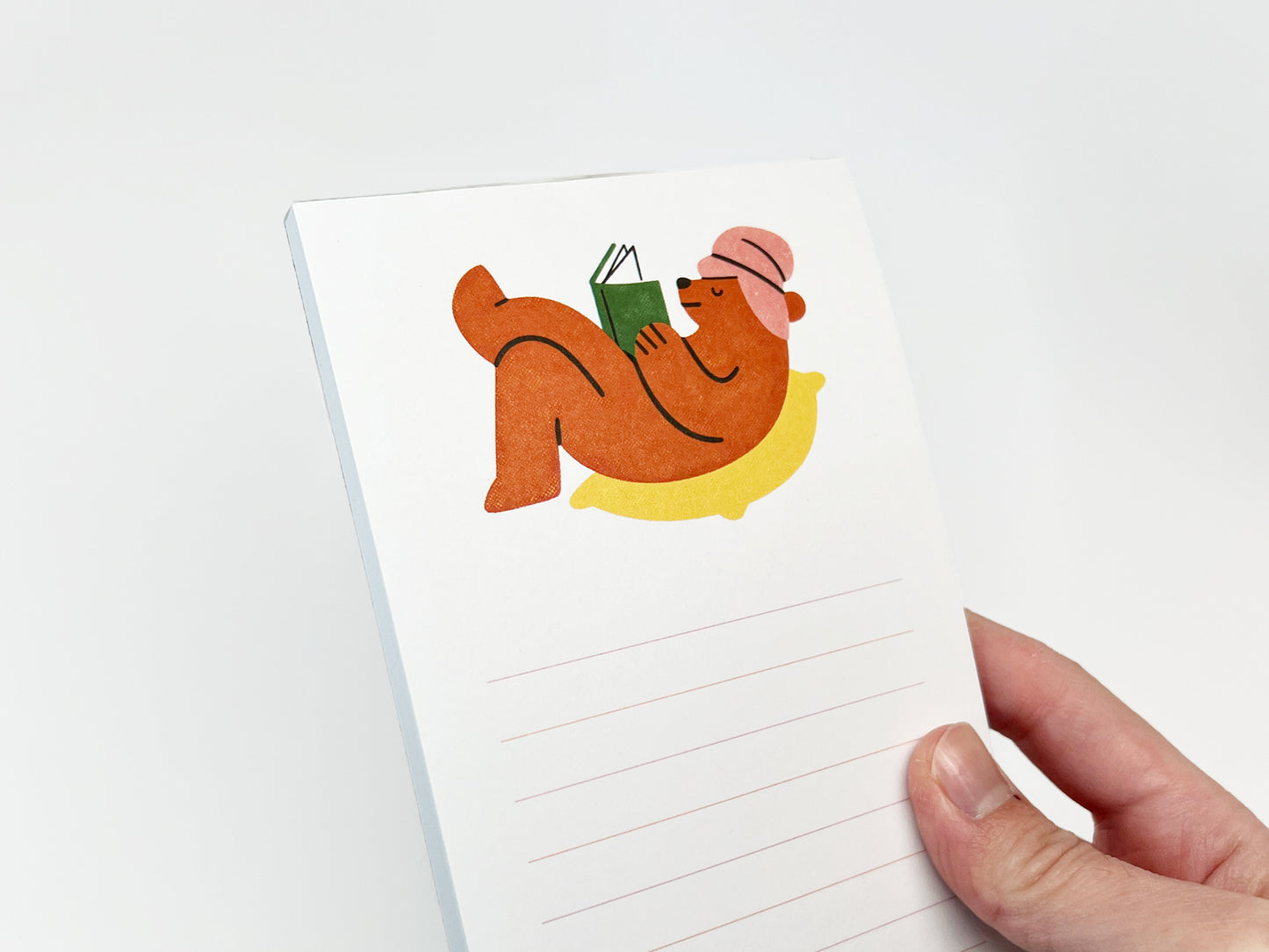 Hand holding a slim vertical lined notepad against a white background, featuring an illustration at the top of a brown bear lounging on a yellow cushion while reading a green book and wearing a pink hat; light pink horizontal lines appear below for writing.
