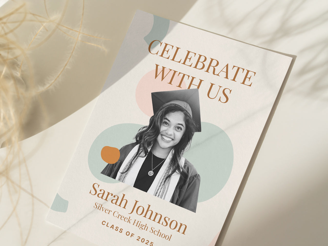 A boho-inspired graduation announcement card with soft pastel abstract shapes and elegant serif text. The 5x7 printed grad invite features a customizable graduate photo and Celebrate With Us in gold-tone typography. The card is displayed in natural lighting with delicate dried flowers, creating a warm, organic aesthetic. This personalized graduation card is perfect for modern, minimalist, and boho-style graduation parties.