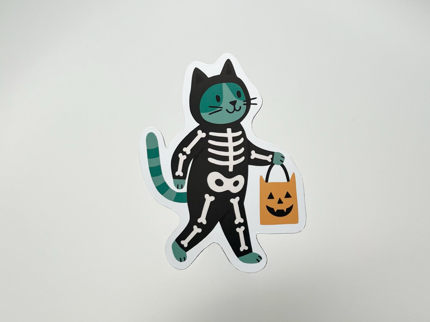 Handmade die-cut Halloween magnet featuring a green and black cat in a skeleton costume, holding an orange pumpkin candy bag. The playful, spooky design is ideal for decorating fridges, lockers, or office spaces and makes a perfect gift for cat lovers and Halloween enthusiasts.