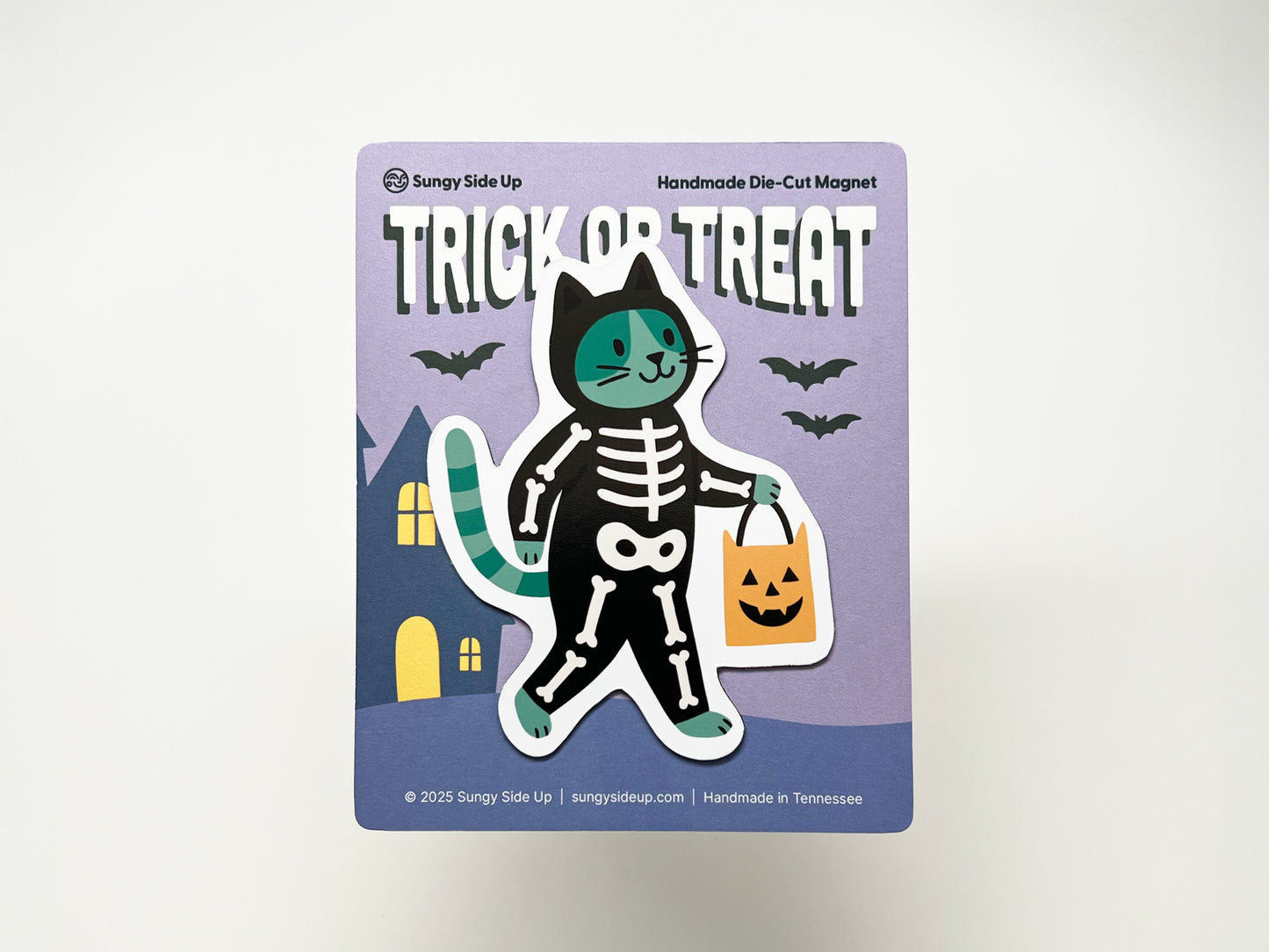 Handmade die-cut Halloween magnet featuring a green and black cat in a skeleton costume holding an orange pumpkin candy bag, displayed on a purple backing card with bats and a haunted house illustration. Perfect for seasonal décor and gifting to cat or Halloween lovers.