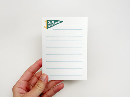 Hand holding a small white lined notepad featuring a green pennant banner that says “Chaos Coordinator” in bold white lettering. The notepad has light blue horizontal writing lines and a clean minimalist layout against a soft white background.