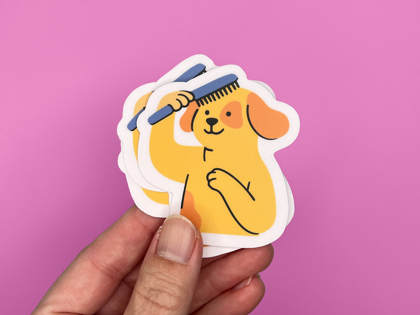 Hand holding a small stack of matte vinyl die cut stickers featuring an illustrated smiling yellow dog brushing its own hair with a blue comb. The dog has floppy orange ears and one paw on its chest. Each sticker has a soft pink border and is shown against a bright pink background, highlighting the layered stickers and compact size.