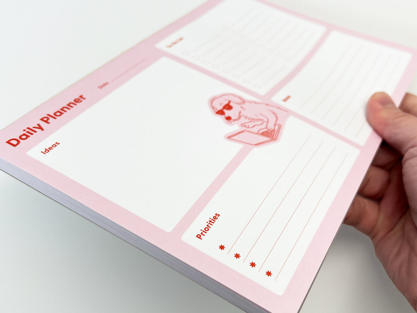 Angled close-up of a pink daily planner notepad being held in one hand, showing multiple stacked tear-off sheets. The layout includes labeled sections for Ideas and Priorities, with star icons marking priority lines. A small illustration of a dog wearing sunglasses and typing on a laptop appears near the center. The background is clean white, highlighting the planner’s design and thickness.