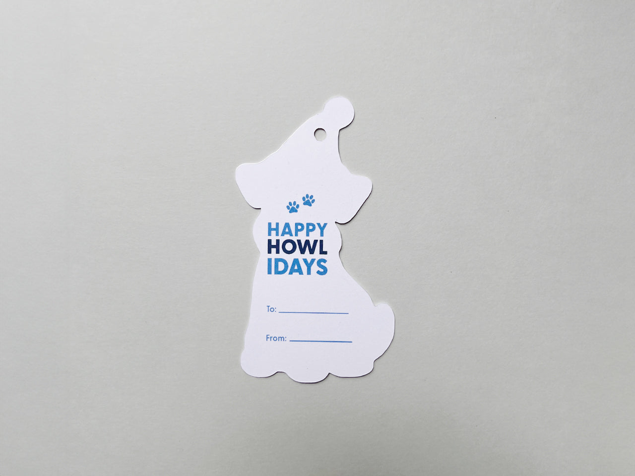Back view of a handmade die-cut Schnauzer gift tag shaped like a dog wearing a winter hat. The tag features the phrase Happy Howlidays in playful blue text with paw print accents and lines for To and From personalization.