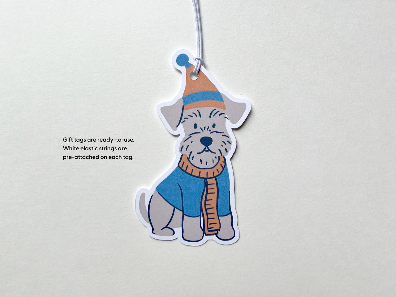 Handmade Schnauzer holiday gift tag with white elastic string pre-attached for easy use. The tag features an illustrated Schnauzer dog wearing a cozy blue and orange winter hat and scarf, ready to add charm to pet lover presents.
