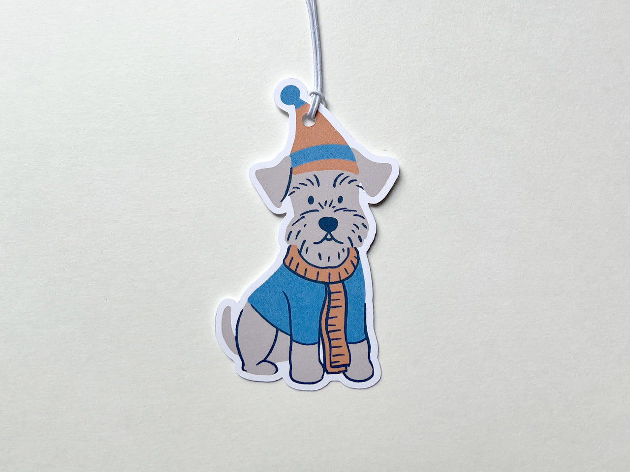 Handmade die-cut Schnauzer holiday gift tag shown with an attached white string for easy tying. The tag features a charming illustration of a Schnauzer dog dressed in a winter hat and scarf, adding a festive and whimsical touch to Christmas gifts.
