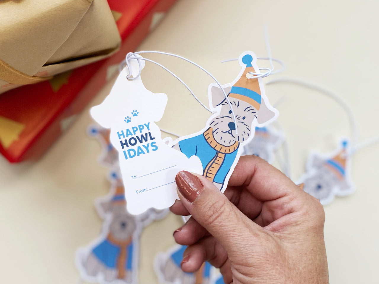 Hand holding a die-cut Schnauzer gift tag set, showing both the illustrated front and Happy Howlidays text on the back with To/From lines. Multiple holiday gift tags with white elastic strings displayed in the background, ready for Christmas gifting.