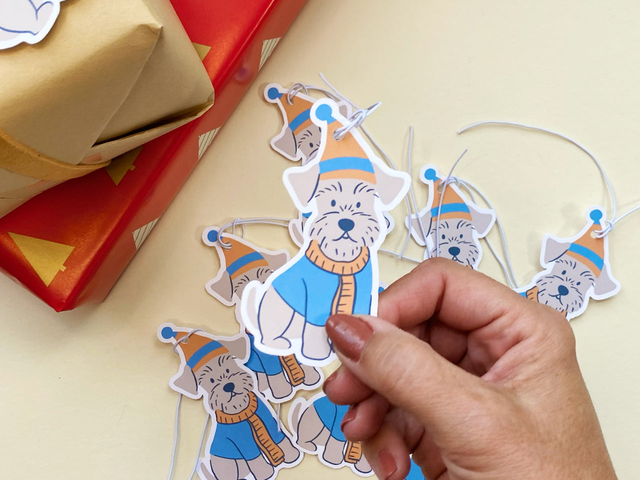 Close-up of a hand holding a handmade Schnauzer gift tag with a blue and orange winter hat and scarf. The background shows a collection of similar die-cut tags with attached white elastic strings, ready for gift wrapping and holiday decor.