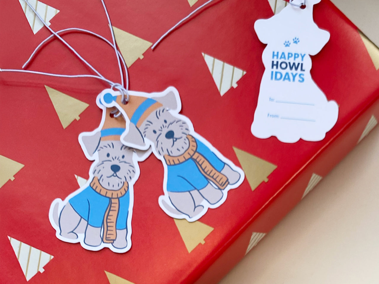 Two handmade Schnauzer gift tags tied with white elastic string rest on red Christmas wrapping paper with gold trees. One tag displays the cute illustrated dog, while the other shows the Happy Howlidays To/From design.