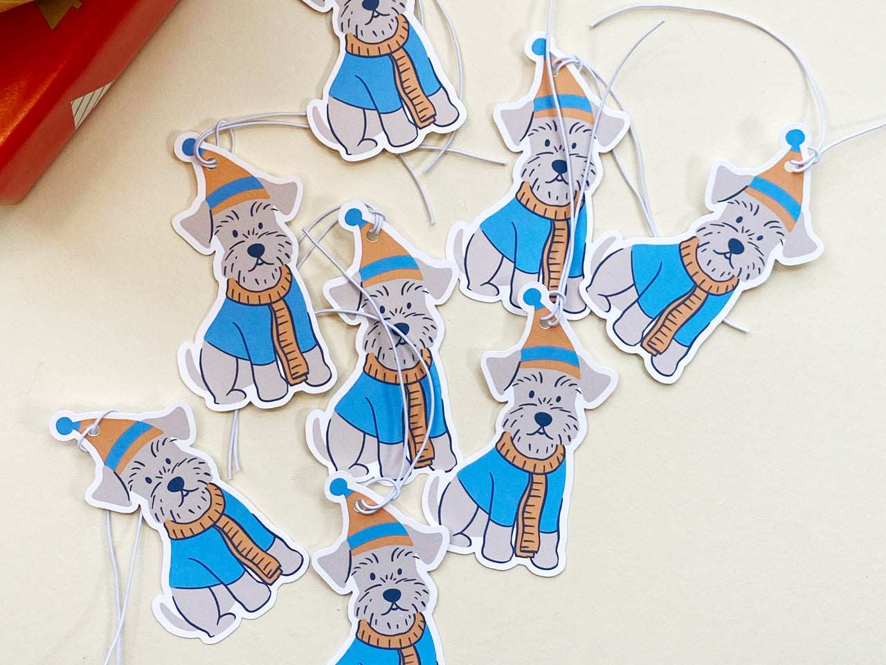 Set of multiple handmade Schnauzer holiday gift tags featuring an illustrated Schnauzer dog wearing a blue and orange winter hat and scarf. Each tag includes pre-attached white elastic strings, making them easy to tie to gifts.