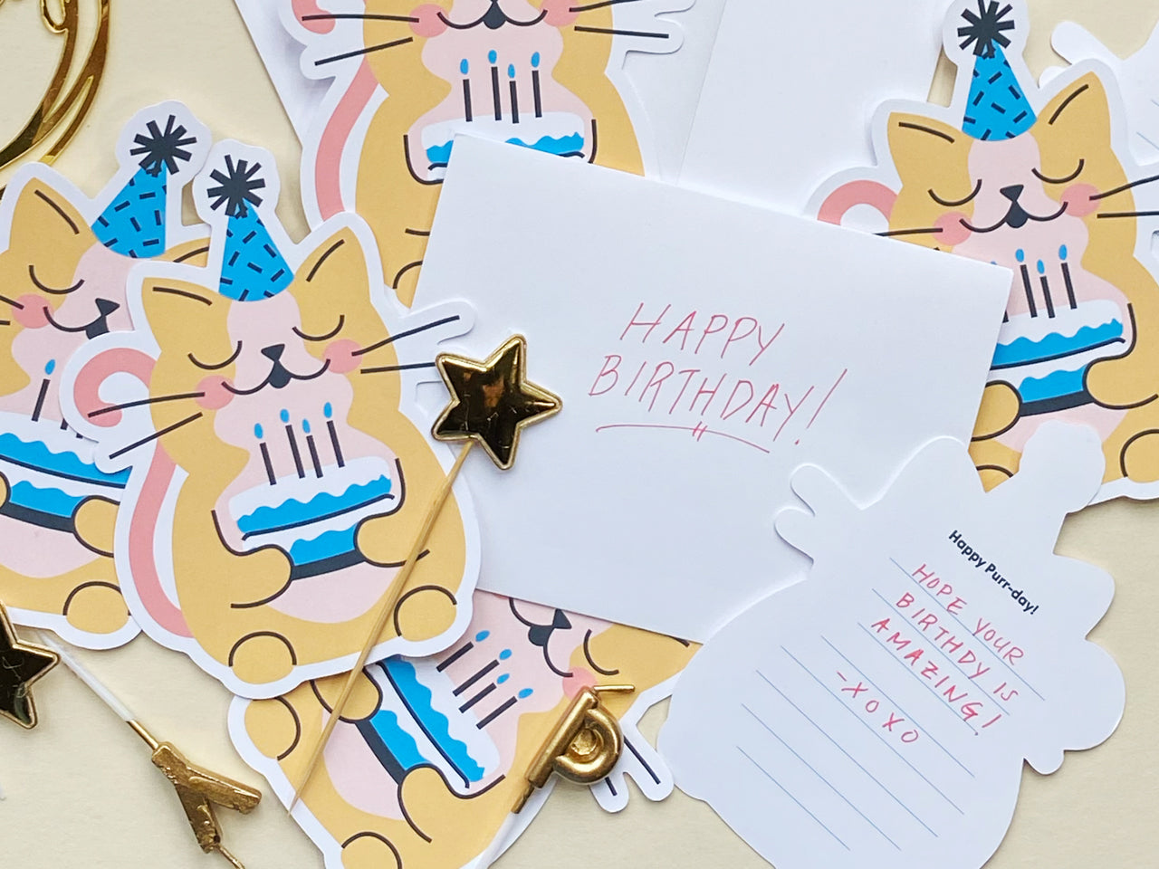 A set of cat-themed birthday cards featuring an illustration of a smiling yellow cat wearing a blue party hat and holding a birthday cake. The background includes several cards, envelopes, gold star cupcake toppers, and a handwritten note in red ink.