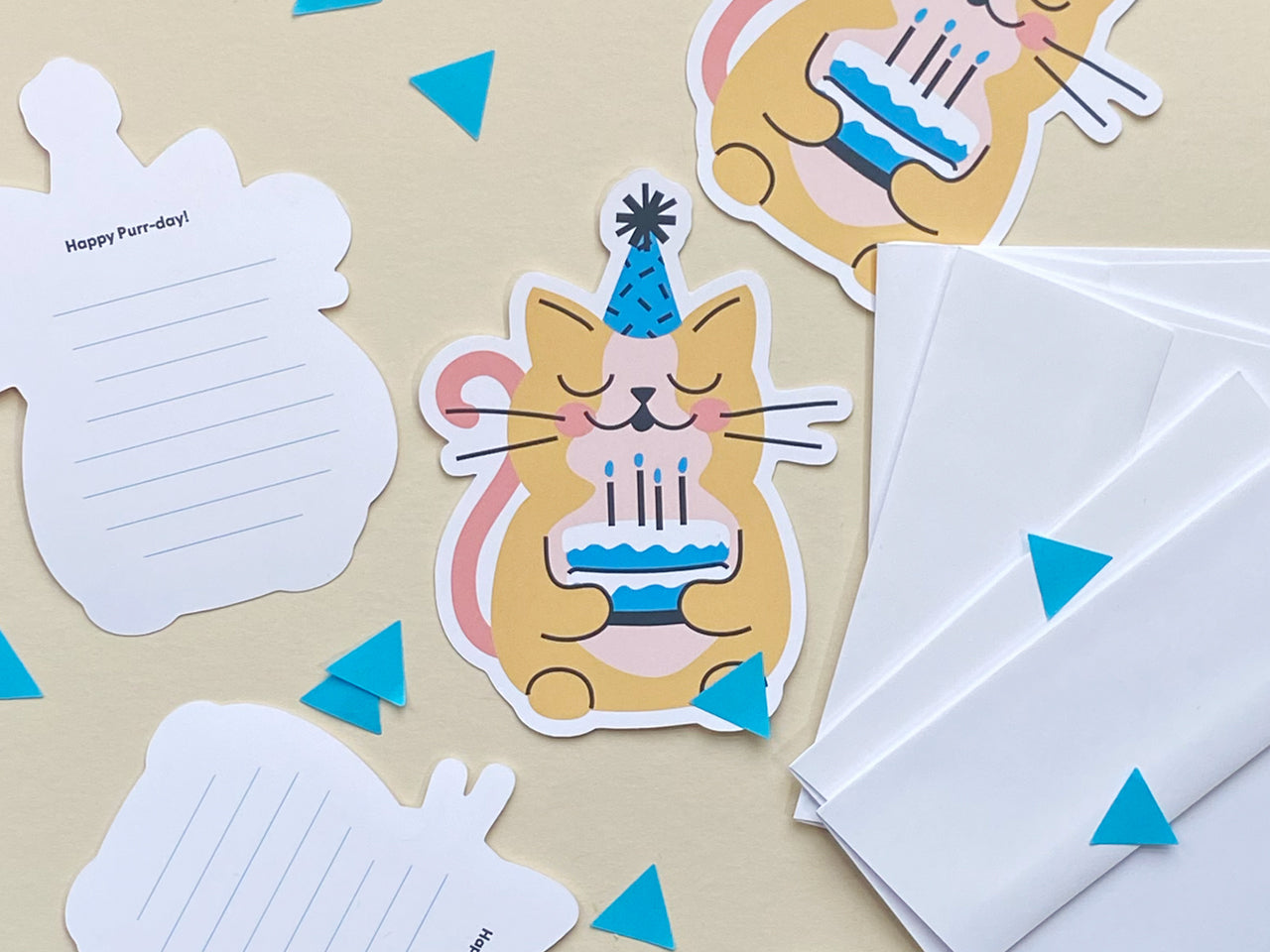 A flat lay display of cat-themed birthday cards featuring a cute yellow cat wearing a blue party hat and holding a blue birthday cake. The cards are arranged with white envelopes and show some of the back of cards which are lined for messages.