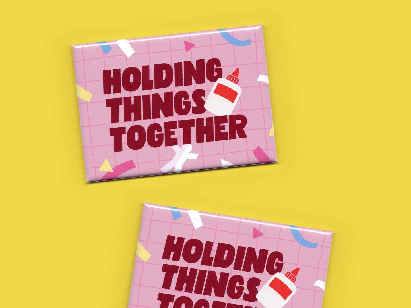 Two pink “Holding Things Together” magnets displayed on a bright yellow background. The magnets feature a pink grid pattern, colorful paper cut out scrap accents, bold dark red lettering, and a small glue bottle illustration.