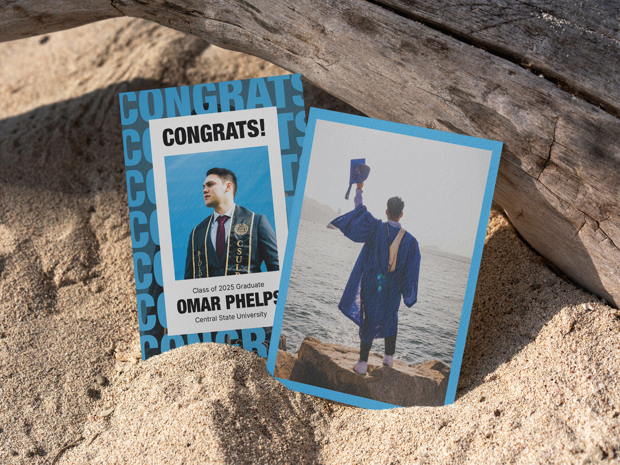 A modern graduation announcement card with bold typography is placed on textured sand, leaning against a piece of driftwood. The card features a customizable photo of a graduate, dressed in a graduation stole, with a congratulatory message in large, bold letters. The back of the card showcases another personalized graduation photo of the graduate wearing a cap and gown. The design uses blue and black text elements, making it a bold, masculine, and modern graduation invitation