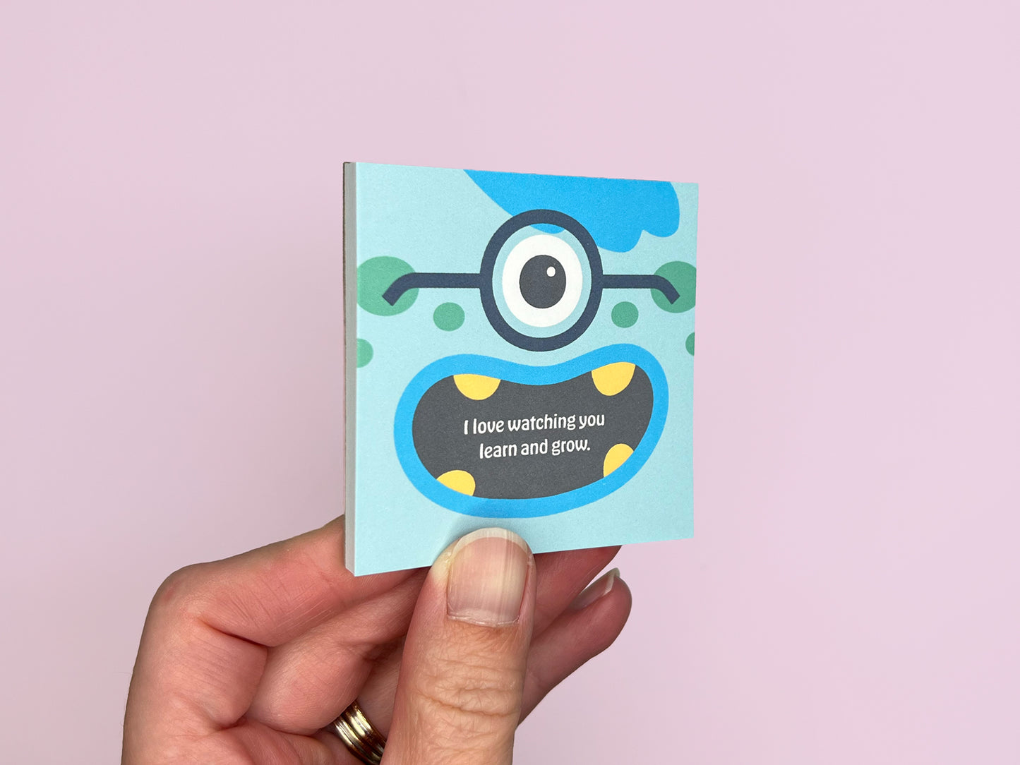 Hand holding a square monster lunch box notes notepad on a soft pink background, showing colorful monster artwork on the front, handmade kids lunchbox notes designed for school lunches and backpacks.