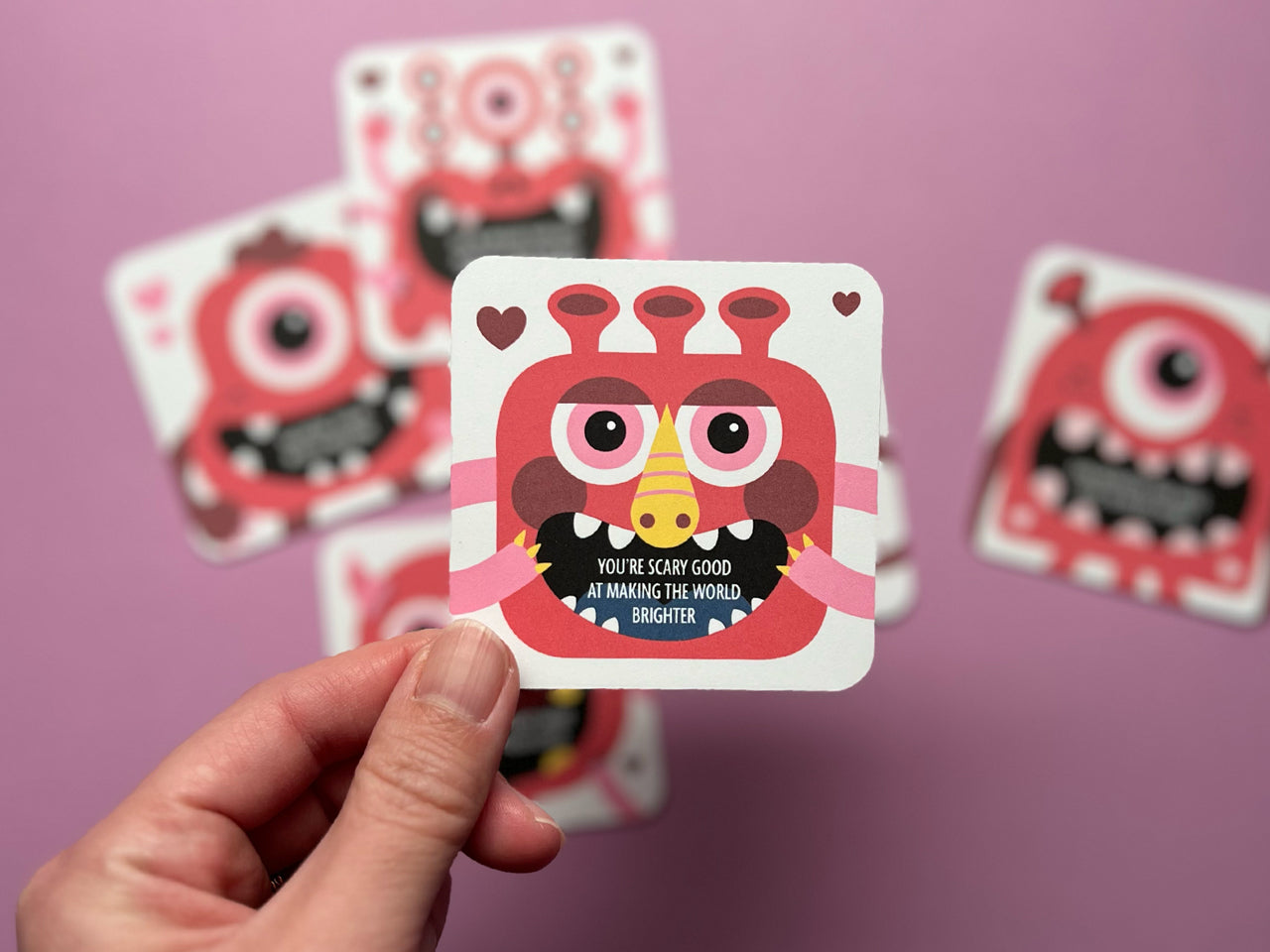 A hand holding a red monster-themed Valentine lunch box note with an encouraging message. In the background, additional cards with playful monster designs and positive messages are slightly blurred, set against a pink background.