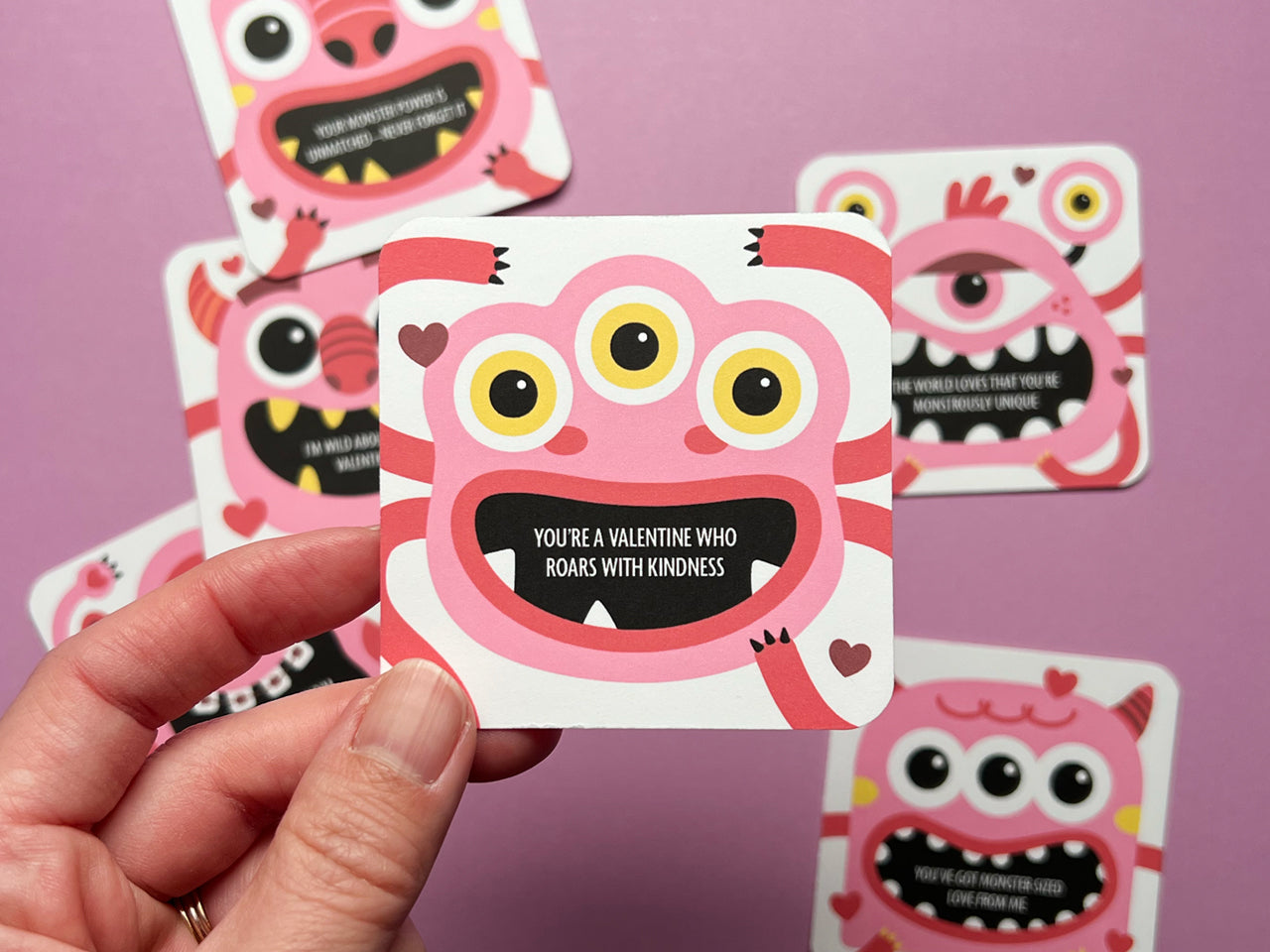 A hand holding a pink and red monster-themed Valentine lunch box note featuring a cheerful three-eyed monster and an encouraging message. In the background, several other colorful and whimsical monster-themed cards are scattered in the background.