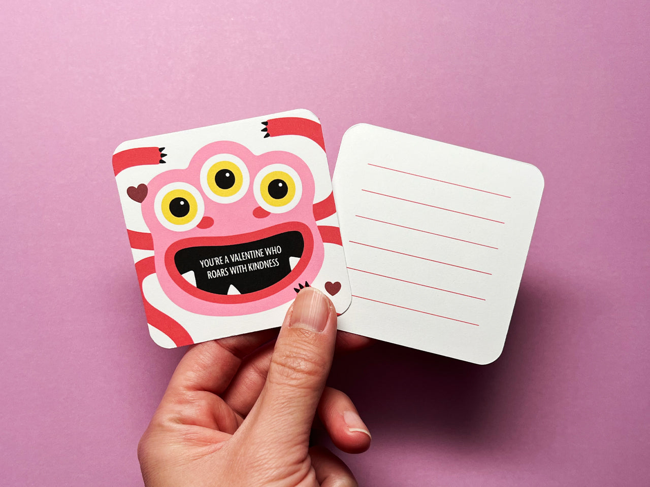 A hand holding a pink and red monster-themed Valentine lunch box note with an encouraging message about kindness on the front. The back of the card includes red lines for writing a personal message, against a pink background.