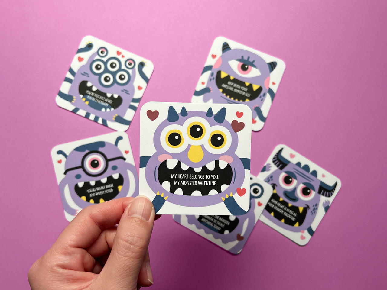A hand holding a purple monster-themed Valentine blank note with encouraging messages on the front surrounded by additional monster-themed lunch box notes with uplifting messages, displayed against a pink background.