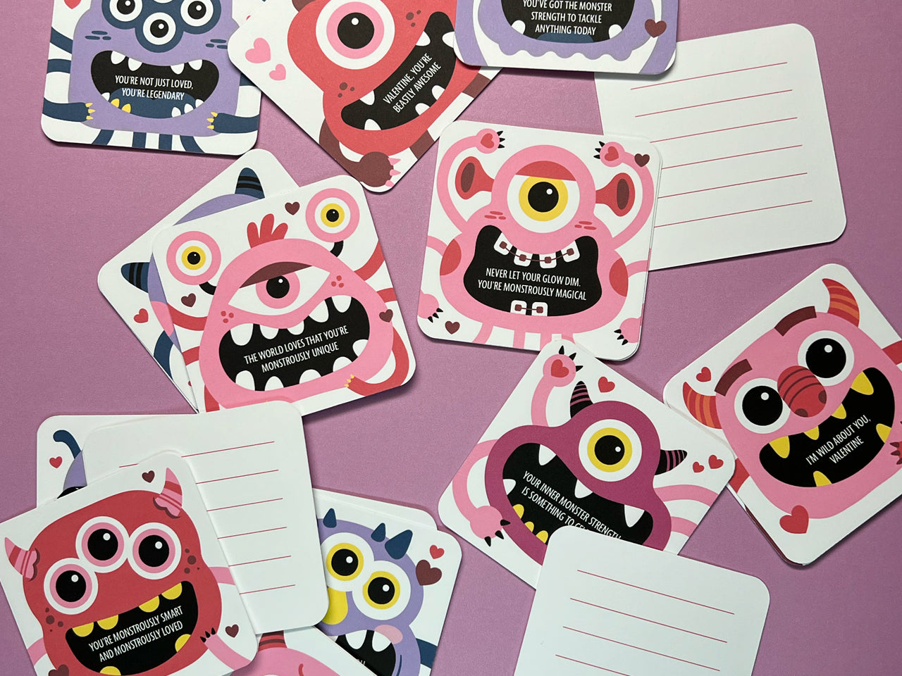 A playful assortment of colorful monster-themed Valentine lunch box notes scattered on a pink background. Each card features a unique design with cheerful monster illustrations and uplifting messages. The backs are lined for personalized notes.
