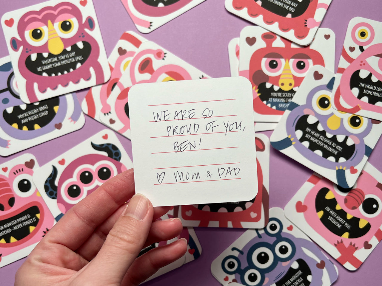 A hand holding a monster-themed Valentine lunch box note with a handwritten message that reads, We are so proud of you, Ben! ♥ Mom & Dad. The back of the card is lined for personalized notes. In the background are other playful monster-themed cards.