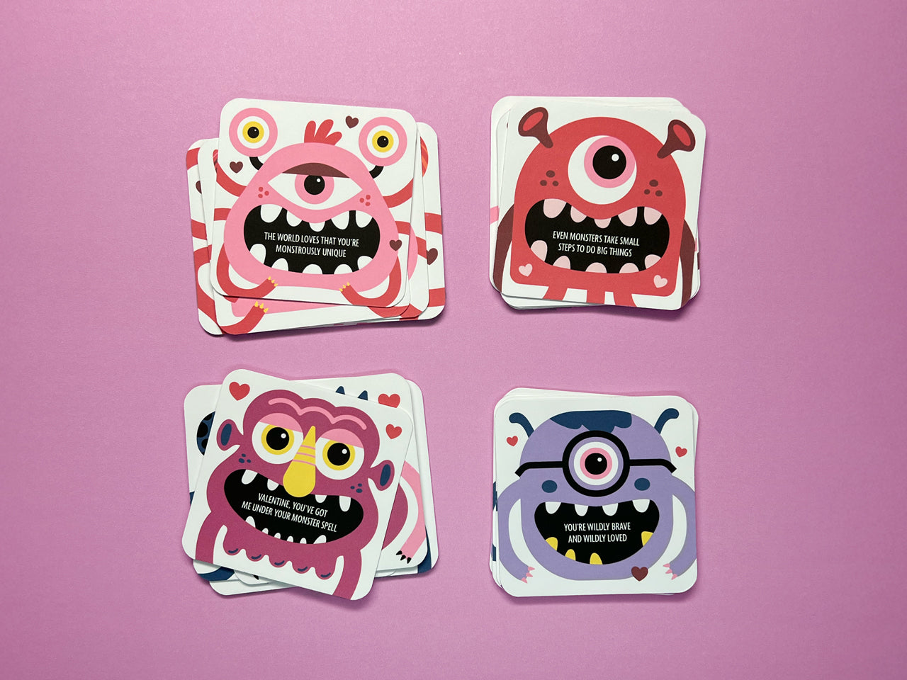 Four stacks of colorful monster-themed Valentine lunch box notes in pink, red, plum, and purple, arranged on a pink background. Each stack features unique designs with colorful monsters and playful, encouraging messages.