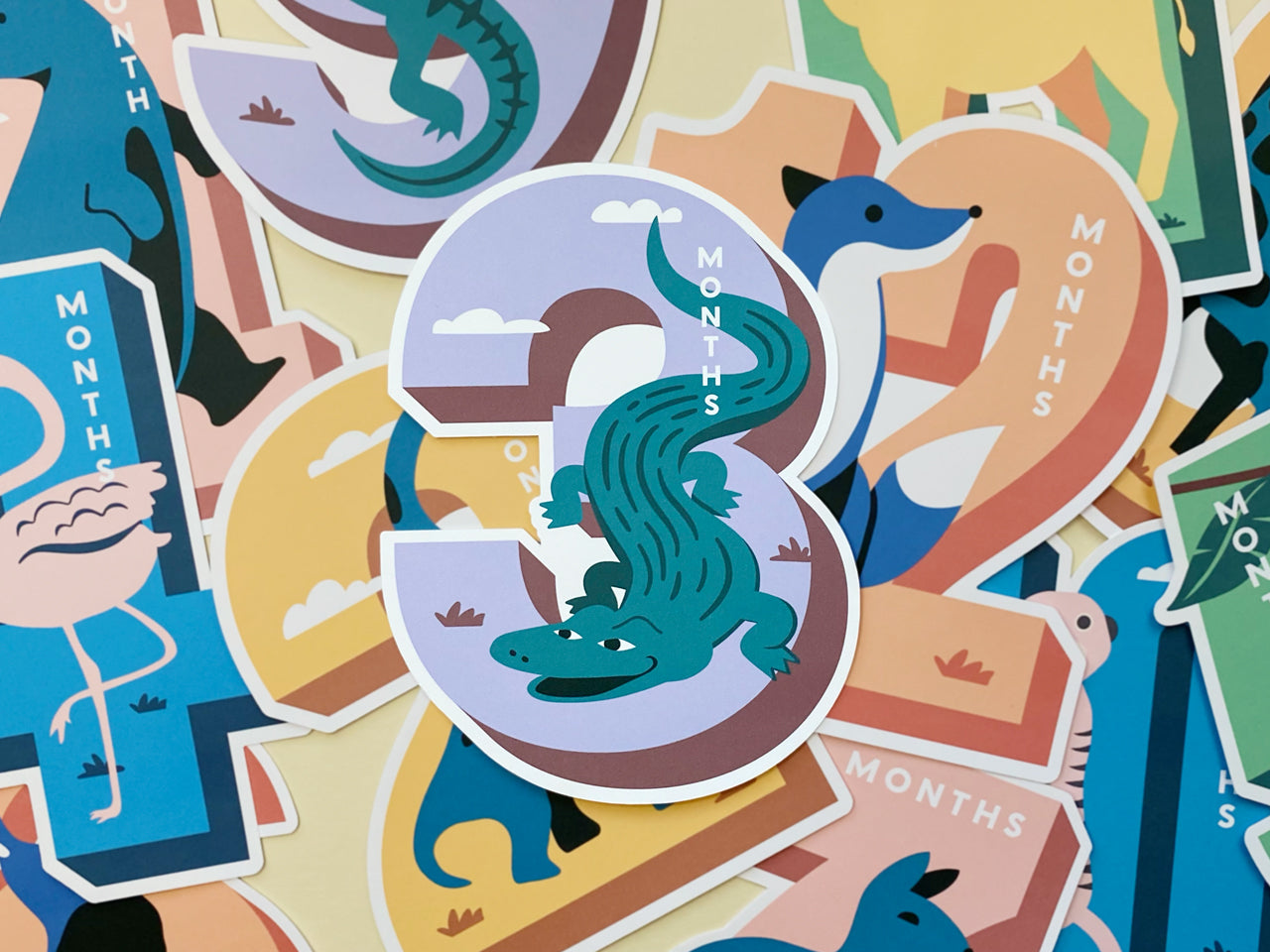 A colorful collection of die-cut milestone cards for baby's first year. The number 3 card in the center showcases a crocodile on a lavender background, surrounded by other milestone cards featuring animals like a flamingo, fox, and camel.