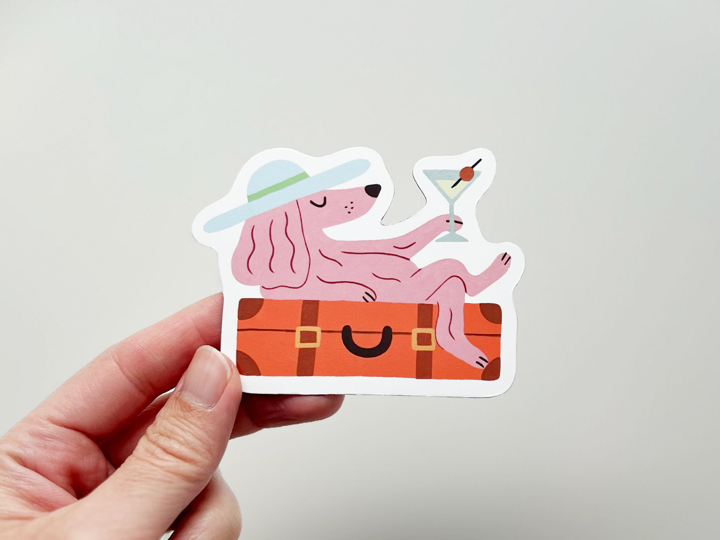 Hand holding a handmade die-cut magnet featuring a pink dog lounging on an orange suitcase while holding a martini with an olive garnish. The dog wears a wide-brim hat with a green band, giving off relaxed vacation vibes. Quirky and fun design for pet lovers and travel enthusiasts.