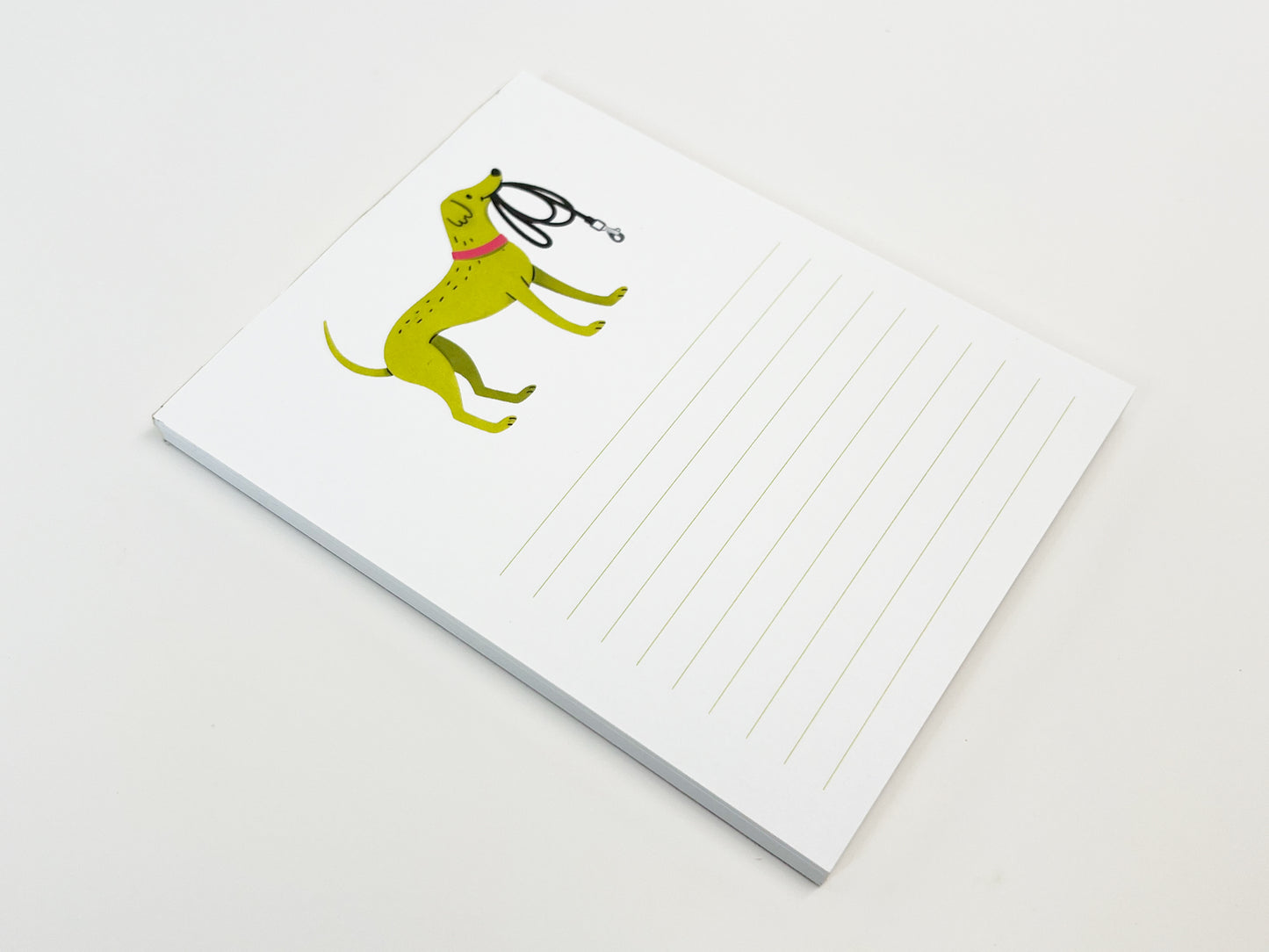A small 4.25 x 5.5 inch lined notepad lying flat on a white surface. The top of the page features an illustrated green dog wearing a pink collar and holding a black leash, with light green horizontal writing lines on the right side of the page and a visible stack of paper sheets beneath.