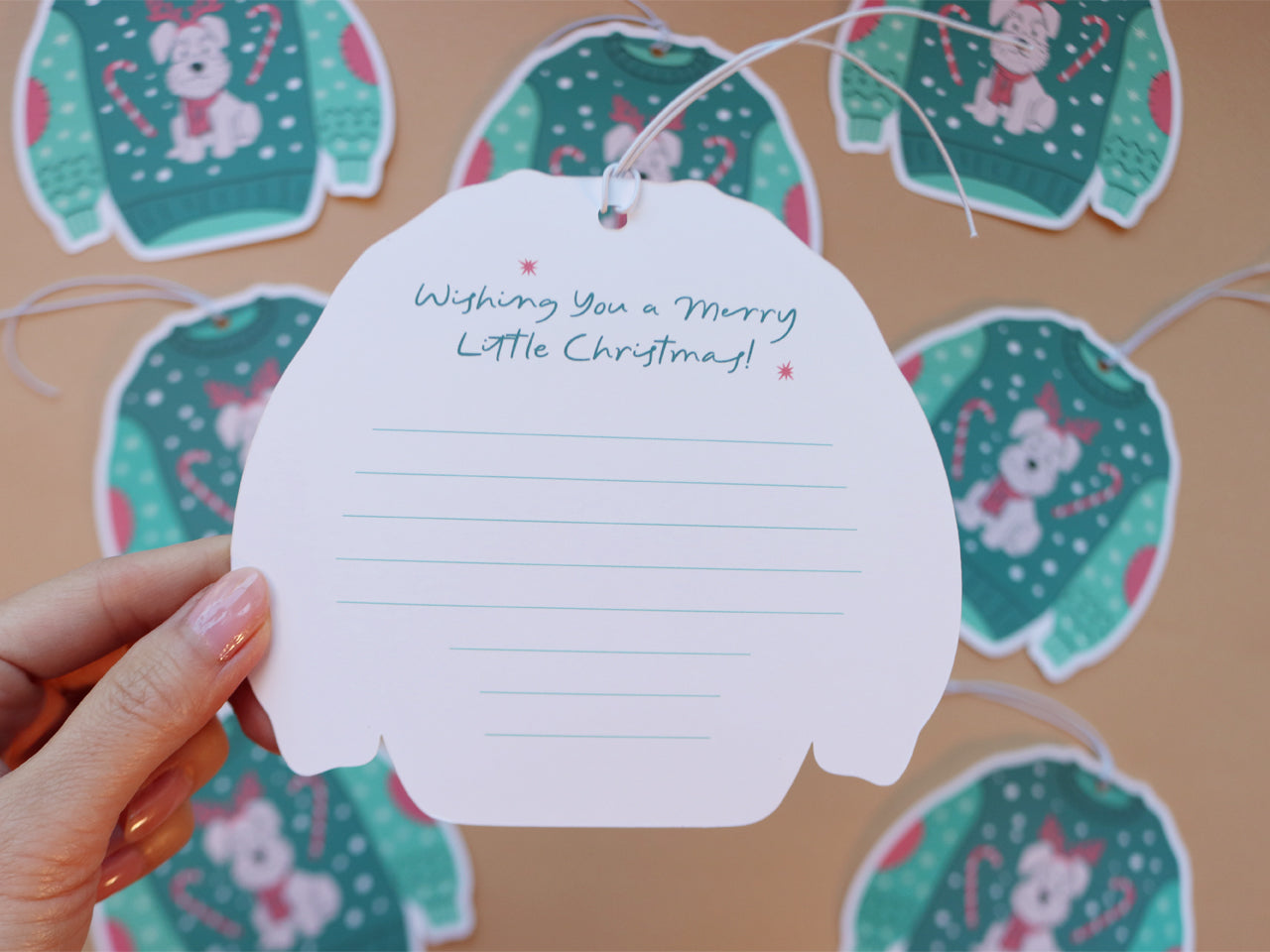 The back of a Christmas sweater tag is held up, showing a space for writing a personal message with the greeting "Wishing You a Merry Little Christmas!" in festive lettering. The tags in the background display the front design with a cute Schnauzer dog and holiday decorations.