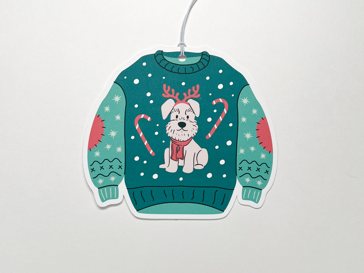 Festive Christmas sweater-shaped gift tag. The sweater is green with playful holiday details, including a cute illustration of a dog wearing reindeer antlers and a red scarf. The sweater is decorated with candy canes and stitched patches on the sleeves, along with snowflakes scattered across the background. A white string is attached to the top for easy attachment to gifts.
