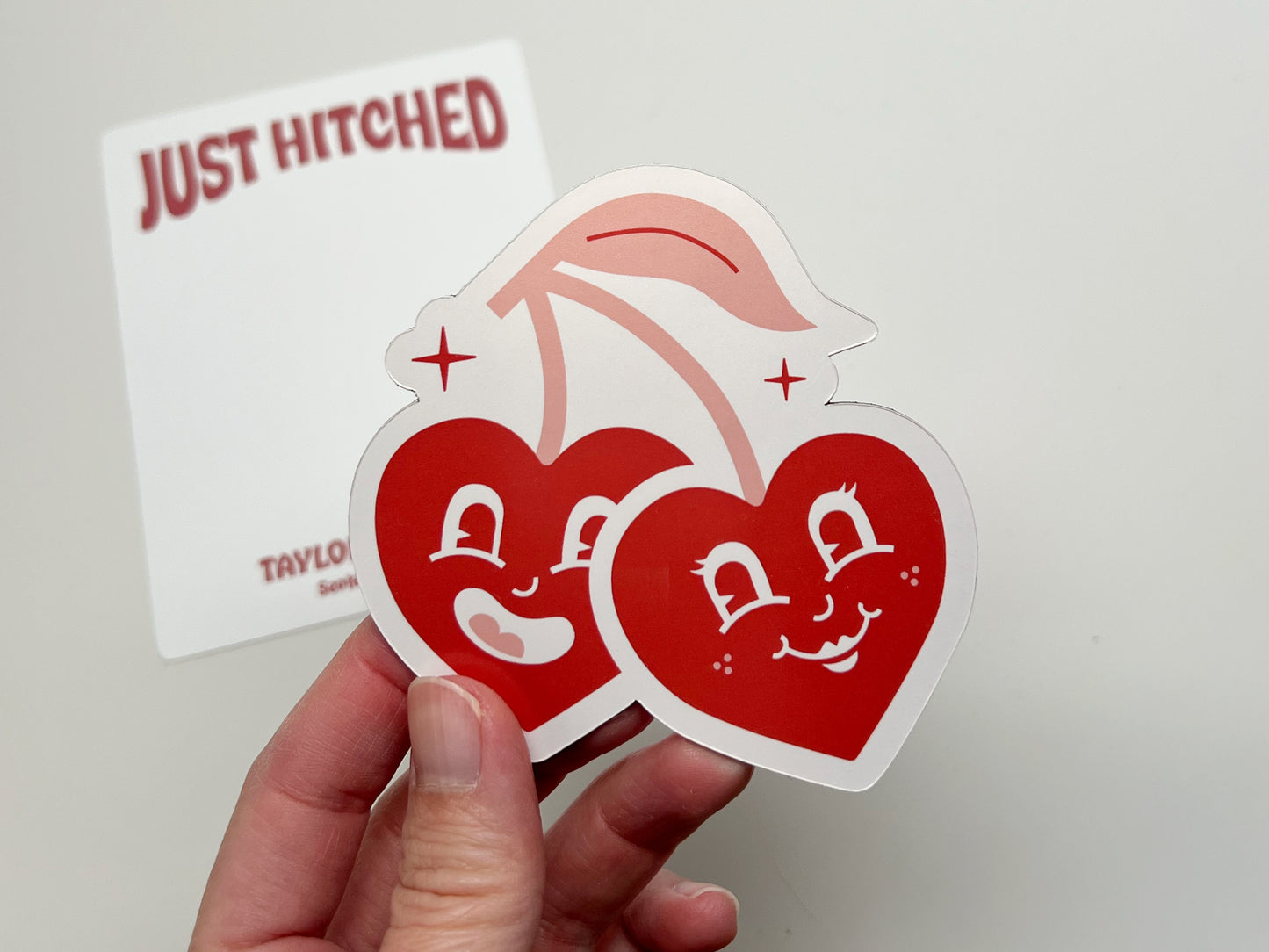 Close-up of a handmade die-cut magnet shaped like two smiling red cherry hearts with retro cartoon faces, held in hand. The backing card in the background reads “Just Hitched,” showing this customizable wedding favor magnet with names and date personalization.