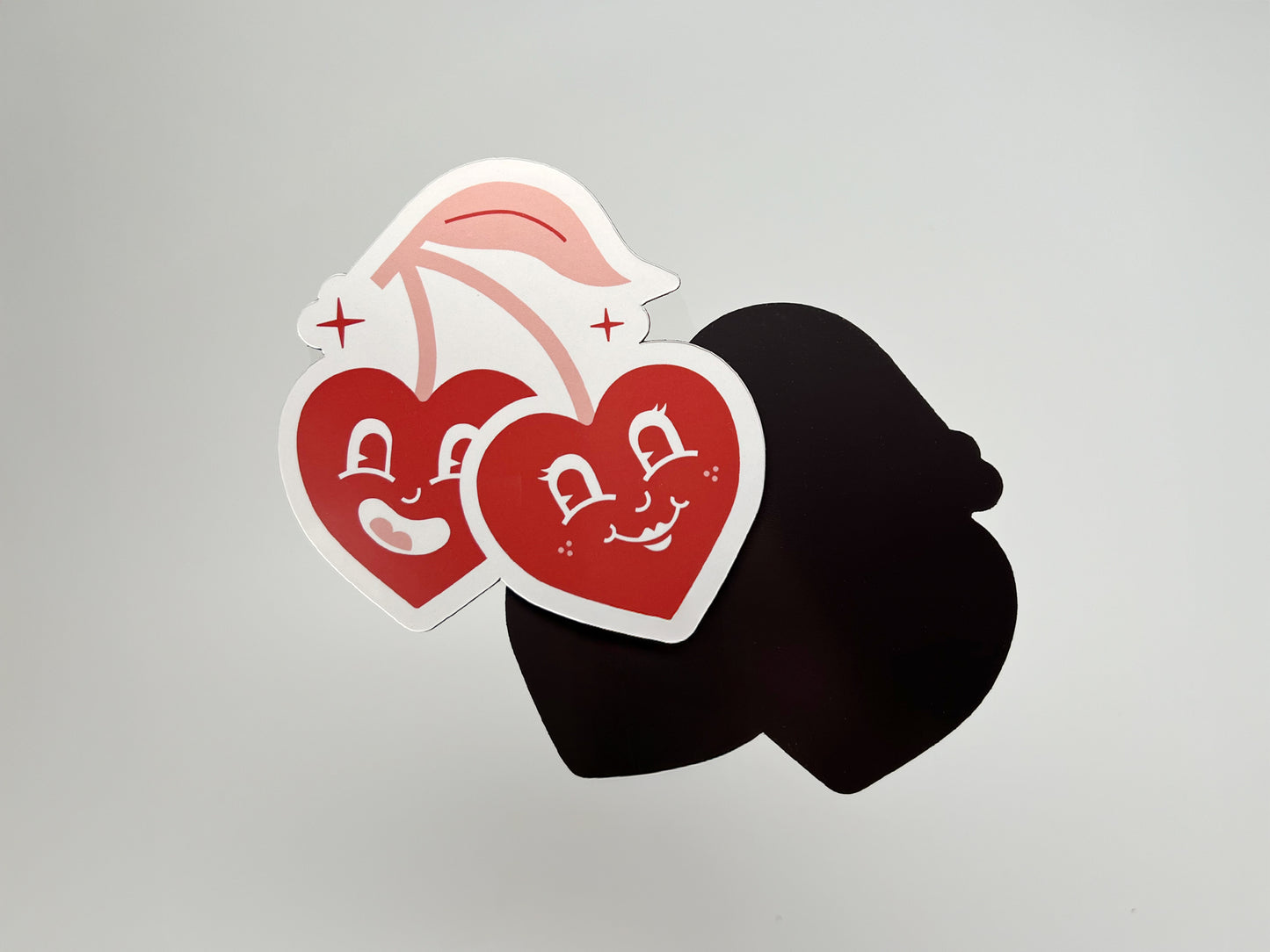 Front and back view of a handmade die-cut magnet shaped like two retro-style smiling cherry hearts with stems. The front features bold red artwork, while the back is solid black magnetic material, showing the quality and durability of this customizable wedding favor.