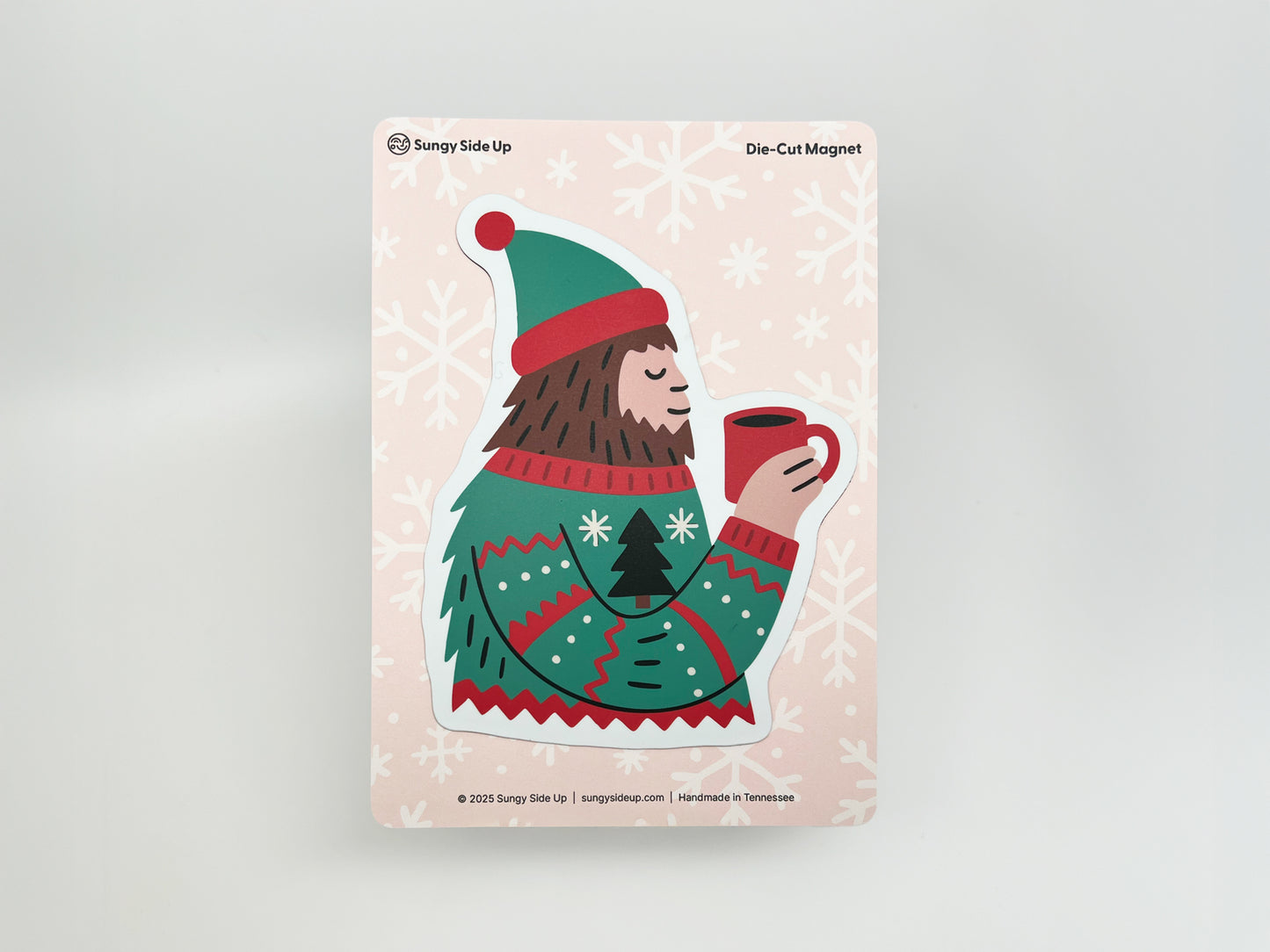 Illustration of a cheerful Sasquatch wearing a green and red winter sweater and matching hat, holding a red mug of hot cocoa with a peaceful smile. The die-cut magnet is displayed on a pink backing card decorated with white snowflakes, branded with “Sungy Side Up” and labeled “Die-Cut Magnet” at the top.