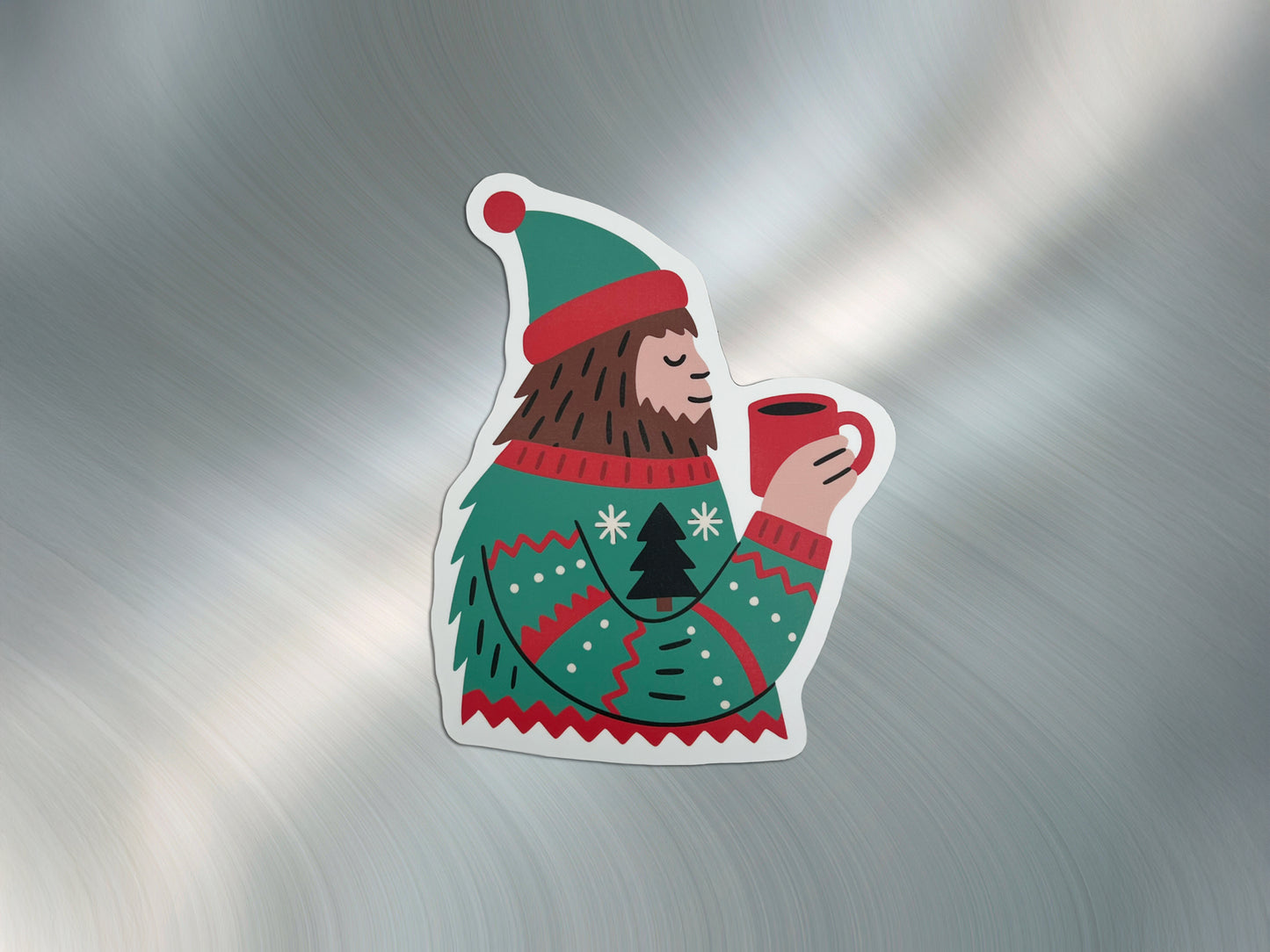 Die-cut magnet featuring a bearded Sasquatch wearing a green and red winter sweater and hat, holding a red mug of hot cocoa. The magnet is displayed on a metallic silver background, highlighting its colorful design and clean white border.