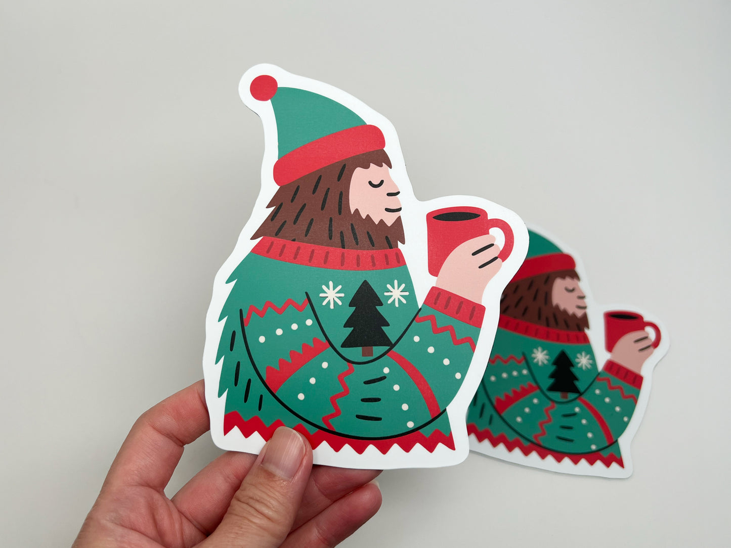 A hand holding a die-cut magnet shaped like a Sasquatch wearing a festive green and red sweater with a Christmas tree design, holding a red mug of hot cocoa. Another identical magnet is slightly blurred in the background against a plain light surface, highlighting the product’s colorful details and glossy finish.