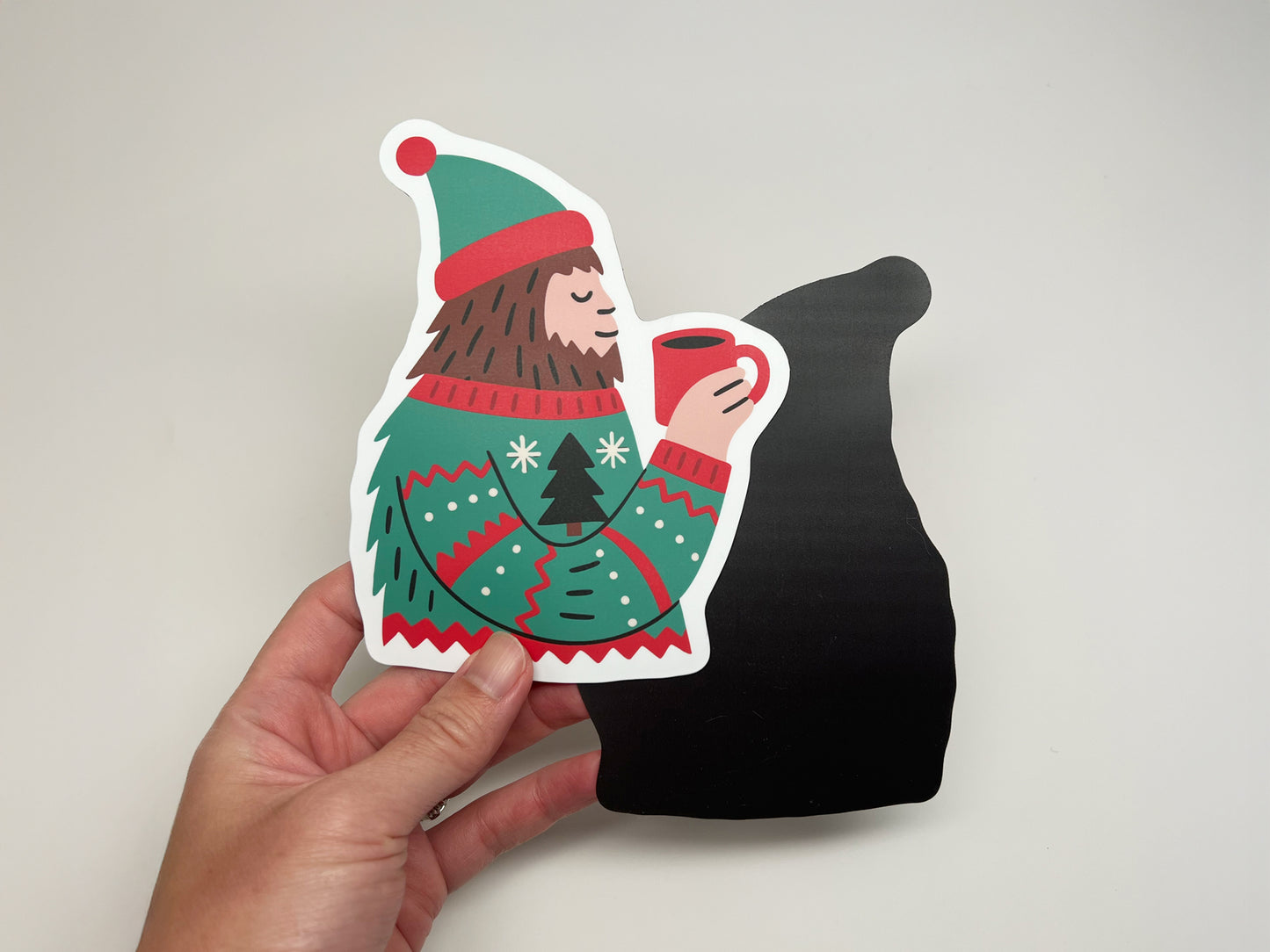 A hand holding a die-cut Sasquatch magnet showing a bearded figure in a red and green winter sweater and hat, holding a red mug of hot cocoa. The front side shows the colorful design, while the black magnetic backing is visible behind it, demonstrating both sides of the magnet. Photographed against a plain light background.