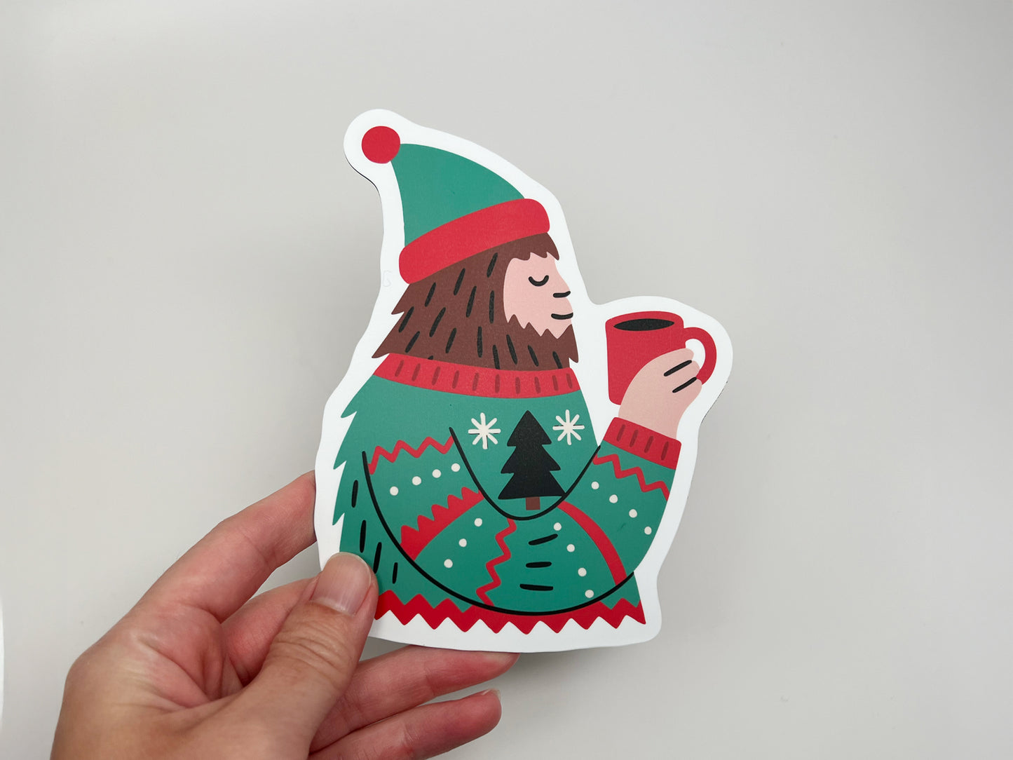 A hand holding a die-cut magnet shaped like a Sasquatch wearing a festive green and red winter sweater and matching hat, holding a red mug of hot cocoa. The magnet features bold colors and clean white edges, shown against a plain light gray background.