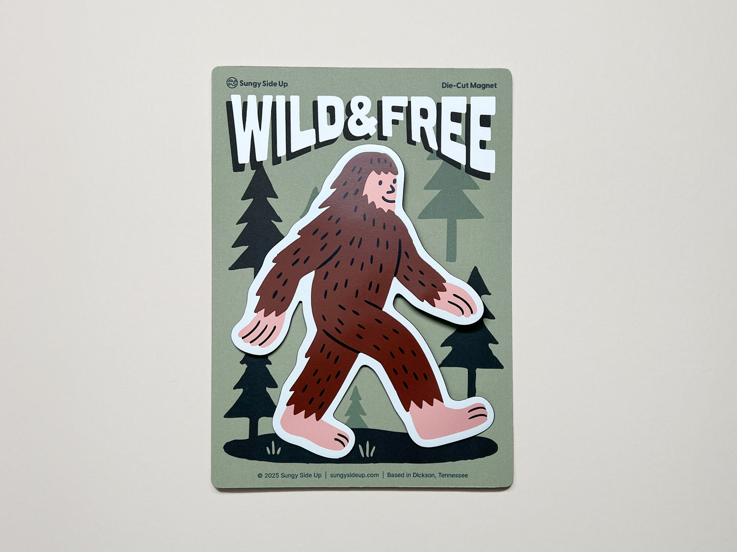 Handmade die-cut magnet featuring a cute brown Sasquatch walking through a forest with the phrase "WILD & FREE" above; quirky Bigfoot fridge magnet by Sungy Side Up, perfect for nature lovers, cryptid fans, and whimsical gift giving.