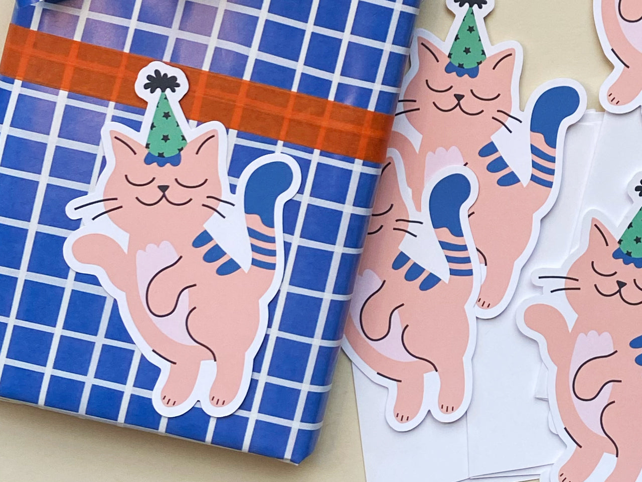 A close-up of several die-cut note cards featuring a pink cat wearing a green party hat with a happy expression and playful pose. A wrapped gift with a blue grid pattern and orange ribbon is placed in the background.