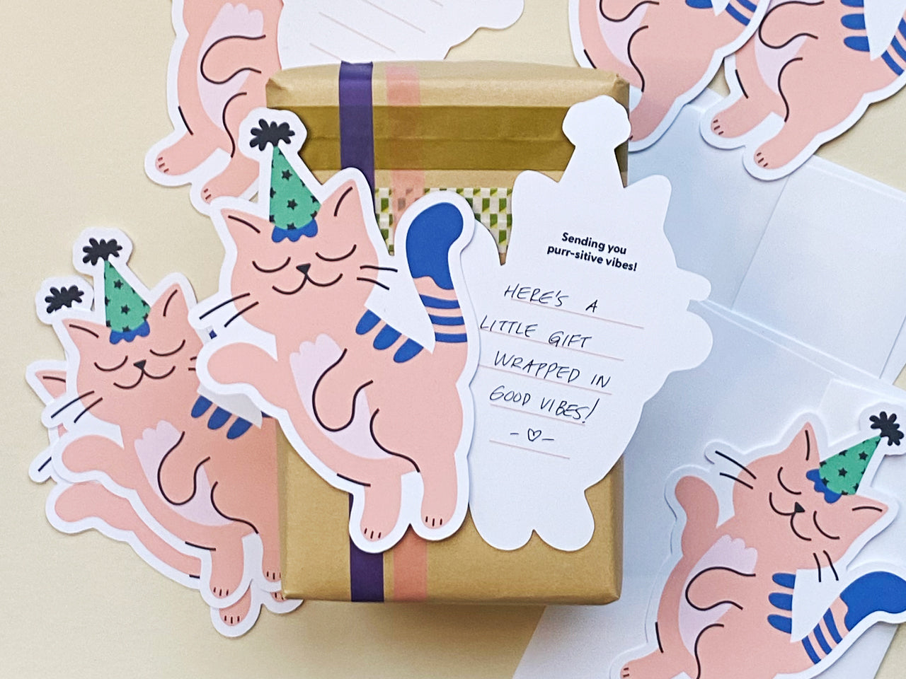 A close-up of a small, gift-wrapped box with brown kraft paper and purple ribbon. A die-cut note card of a pink cat in a green party hat is attached, and the back of the card shows a handwritten message.