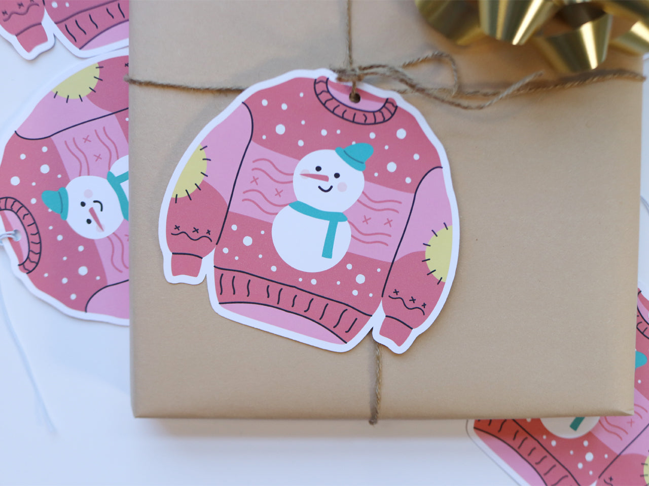 A Christmas sweater-shaped gift tag featuring a whimsical pink design with a snowman wearing a teal scarf and hat. The sweater has yellow patches on the elbows and is decorated with snowflakes. The tag is attached to a wrapped gift with twine, adding a playful touch to the holiday present.
