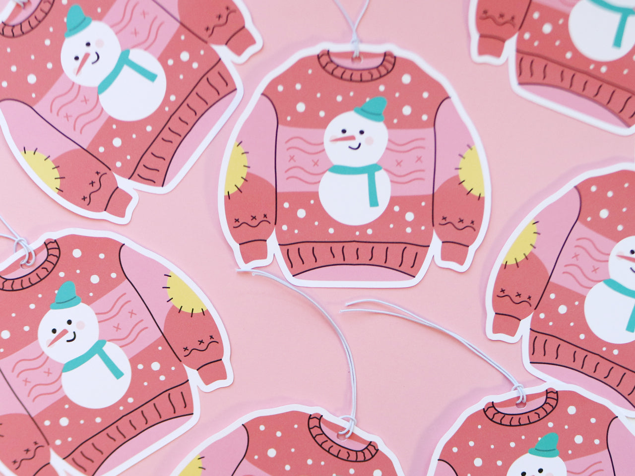 Several Christmas sweater-shaped gift tags, each with a pink background featuring a cheerful snowman with a teal hat and scarf. The sweaters have yellow elbow patches and snowflake designs, all set against a pink surface, adding to the festive and playful feel.
