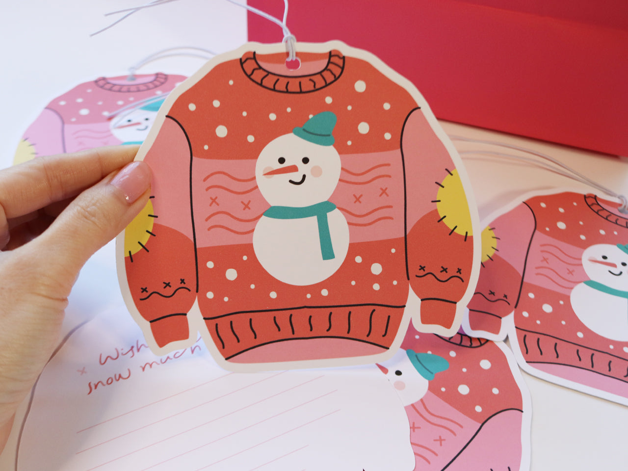 A close-up of a hand holding a pink Christmas sweater-shaped gift tag featuring a snowman with a teal hat and scarf. The festive design includes snowflakes and yellow elbow patches, perfect for adding cheer to holiday presents.