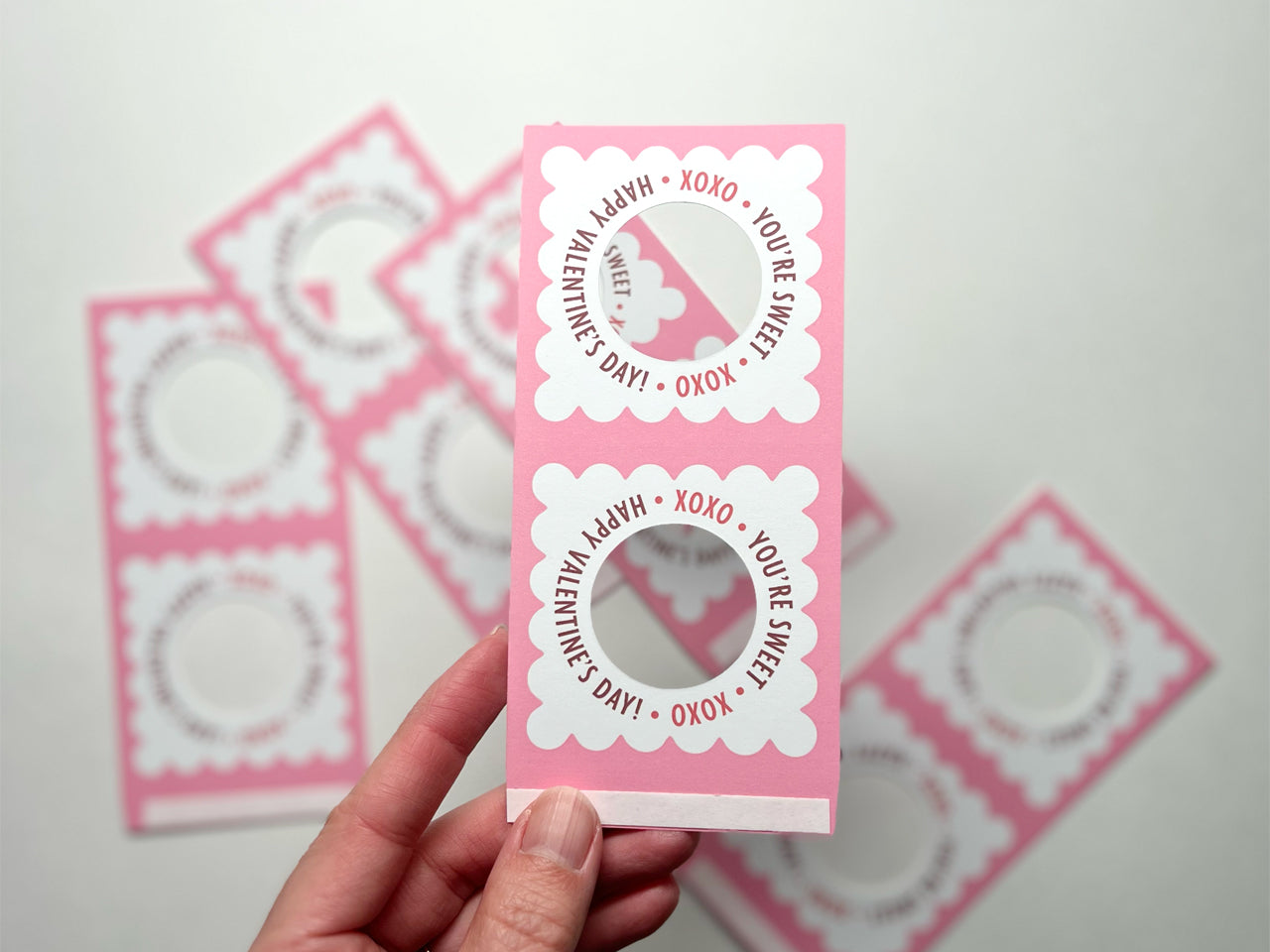 A hand holding a pair of unassembled pink Valentines Day lollipop holders, each featuring a scalloped design and circular cutout for a lollipop. The text reads, XOXO You're Sweet Happy Valentine's Day! More holders are scattered in the background.