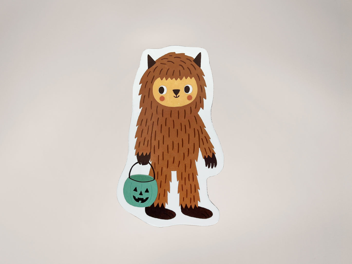 Handmade die-cut Bigfoot Halloween magnet featuring a cute brown woodland creature with a friendly face, holding a teal jack-o’-lantern candy bucket. Quirky cryptid fridge décor perfect for spooky season, trick-or-treat lovers, and unique Halloween gifts.